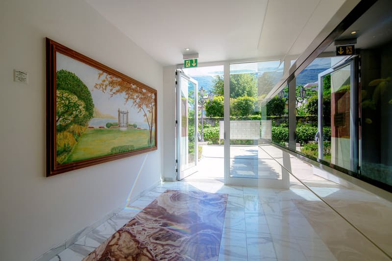 Spacious lobby with large windows overlooking a garden, ornate framed painting on the wall, marble flooring, and glass doors leading to an outdoor terrace or balcony area.