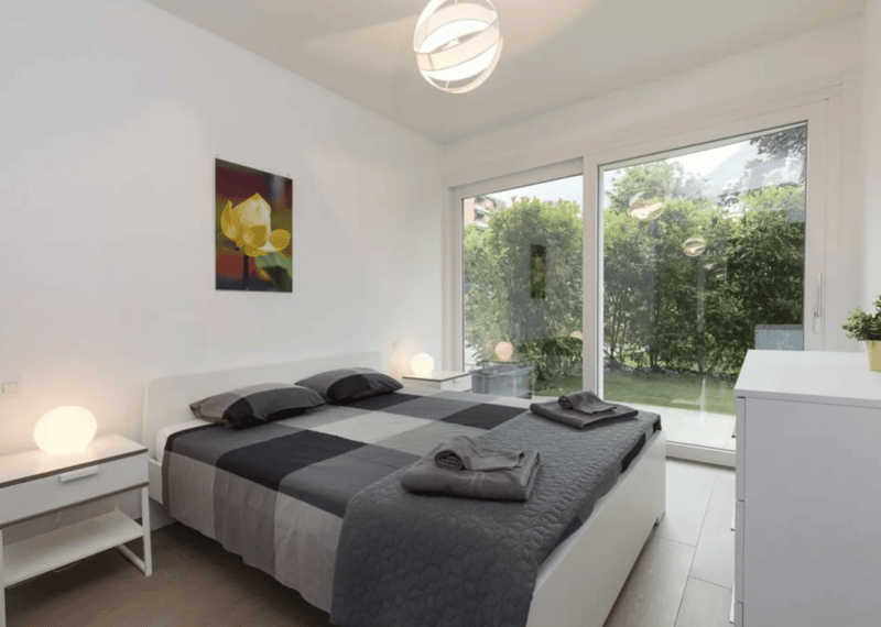 Bedroom with white walls, gray bedspread, nightstands, ceiling lamp, sliding glass door with garden view