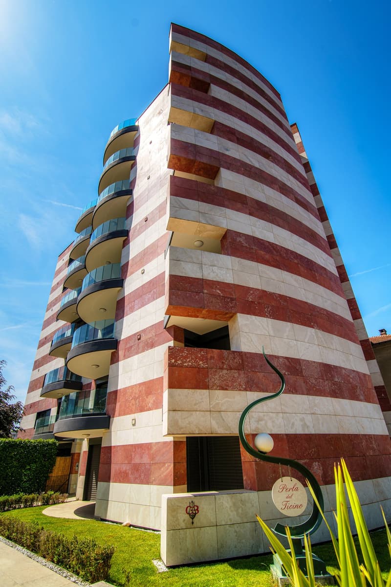 Tall, multi-story building with a striped facade in shades of red, yellow, and white. The building has balconies on each floor and a sign that says 'Perla del Ticino' at the entrance.