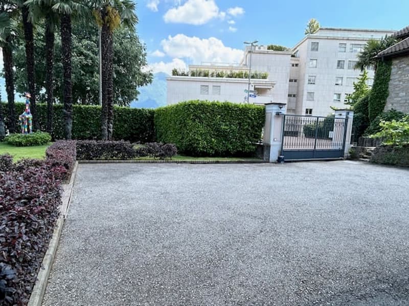 Multi-story apartment building with a gated entrance, surrounded by palm trees and landscaping. The building has a modern, minimalist design with a gray exterior and large windows.