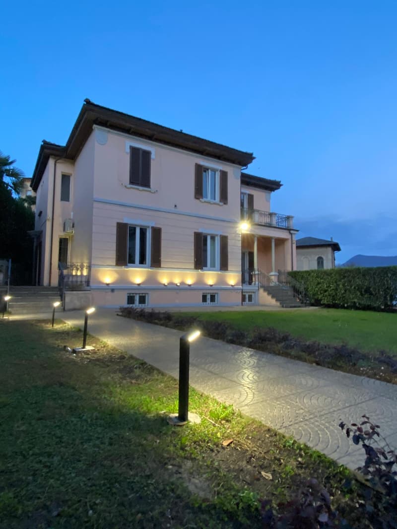 2-story house with a light-colored exterior, balconies, and outdoor lighting illuminating the landscaped garden and walkway in the foreground.