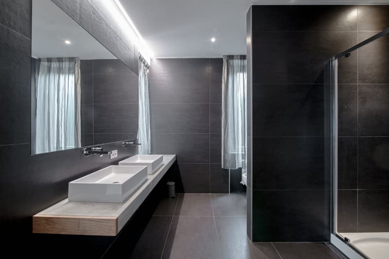 Large modern bathroom with dark gray tile walls, double vanity with white sinks, wooden countertop, and a glass shower enclosure