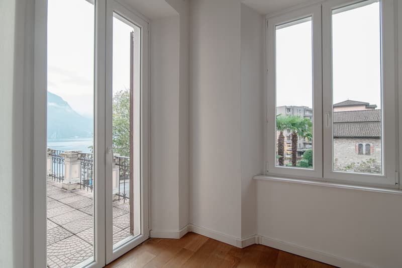 Spacious room with large windows overlooking a balcony and a scenic view of the surrounding buildings and palm trees. The room has hardwood floors and white walls, creating a bright and airy atmosphere.