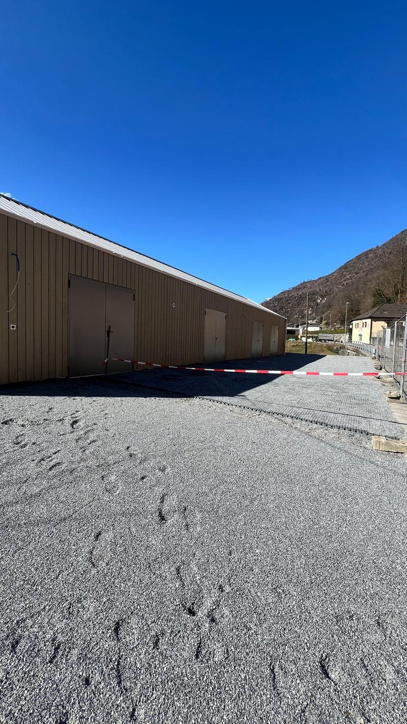The image shows a large brown industrial-style building with a gravel parking area in the foreground. The building has a flat roof and multiple garage doors. The surrounding area appears to be mountainous with a clear blue sky overhead.