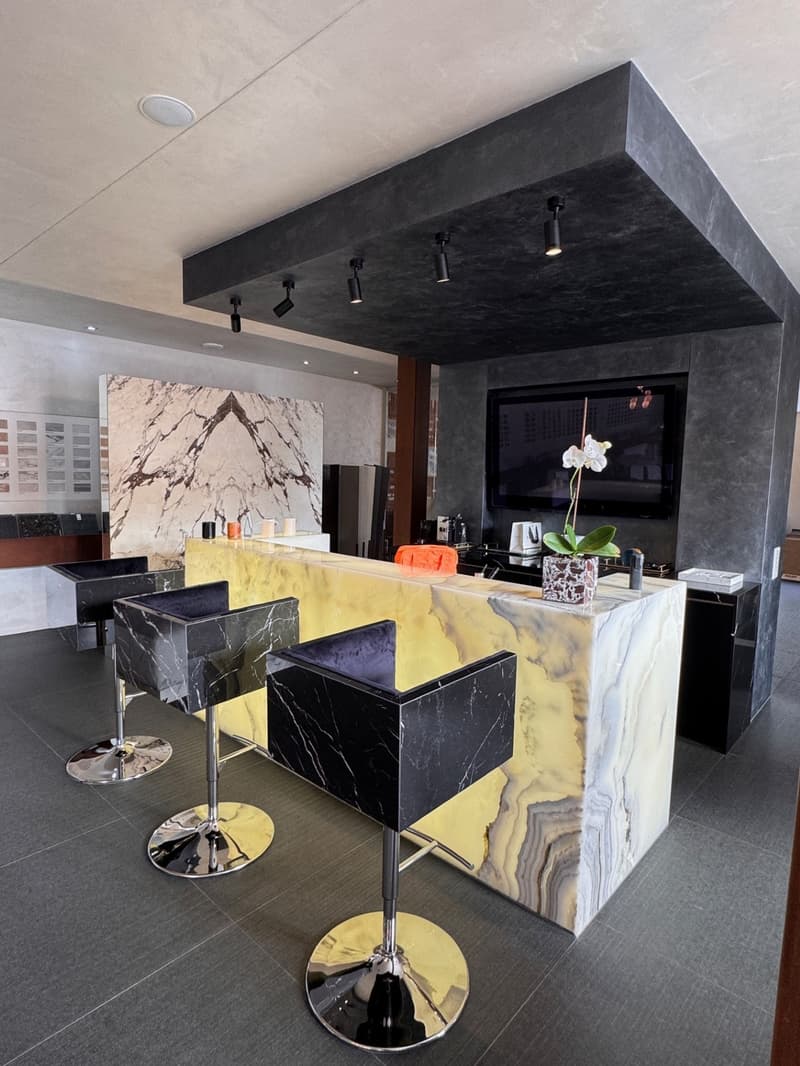 A modern bar counter in the center of the room, with marble countertop, backsplash, black chairs, TV mounted on the wall, flower vase on the counter, and wall sample in the background.