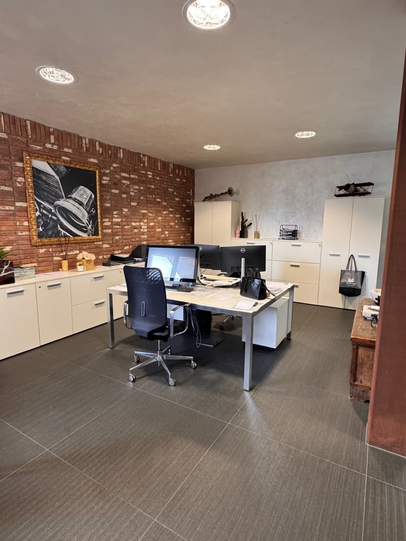 An office with a brick wall, desks, computer monitors, cabinets, a chair, and a picture frame