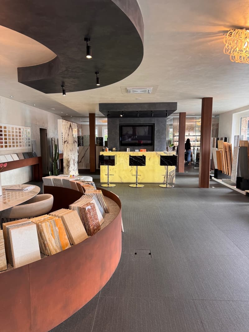 A large open space with a circular display of tile samples, a yellow countertop, stools, and a television. The ceiling features a curved design with multiple ceiling lights and a chandelier.