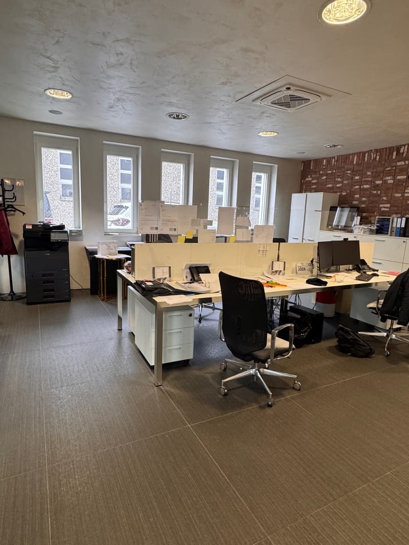 Empty office with desks, chairs, and office equipment. Windows with white frames, brick wall with cabinets, ceiling lights.