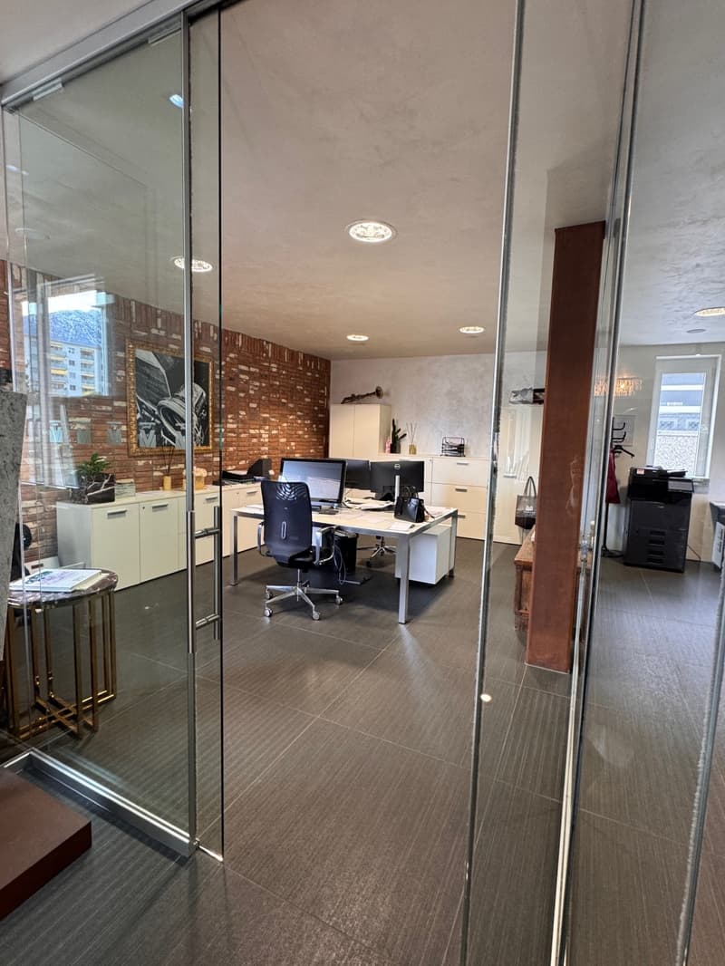 Modern office space with glass walls, red brick wall, desks, monitors, chairs, printer, cabinets, potted plants, tiled floor, lamps