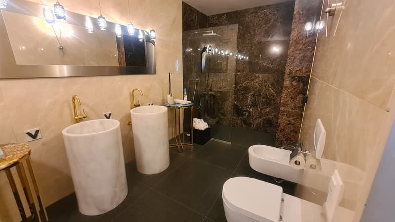 Two freestanding marble sinks, shower stall, toilet, bidet, marble walls, modern lighting fixtures