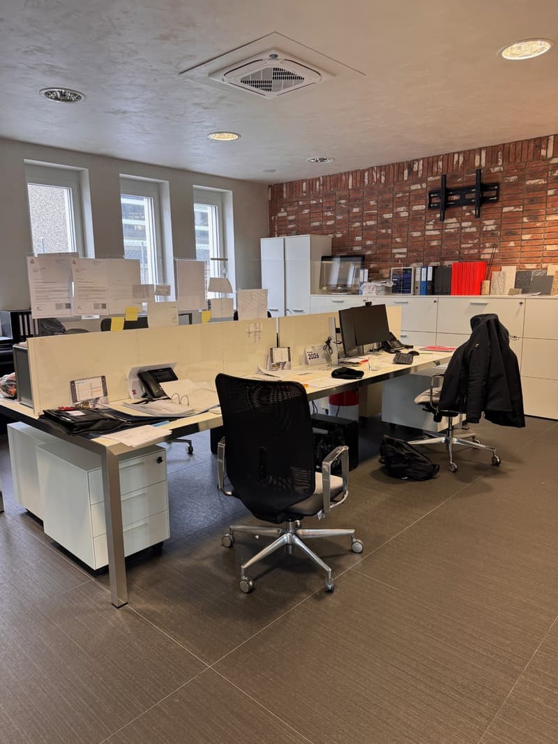 Modern office space with desks, computer monitors, office chairs, cabinets, and a red brick wall.
