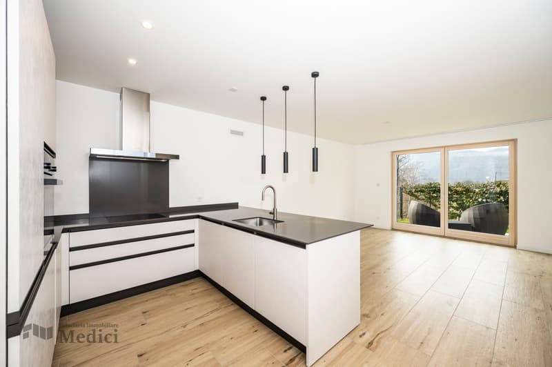 Modern kitchen with a large central island, stainless steel hood, sink with faucet, black countertops, white cabinets, three hanging lamps, wooden floors, large glass doors to a balcony