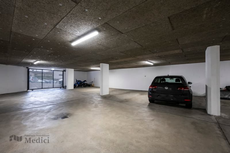 A spacious parking lot with a car parked, an enclosed gate, motorcycles, and ceiling lights