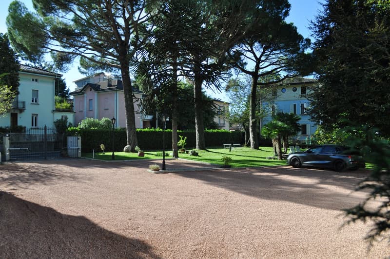 pebbled driveway, gated entrance, parked car, lawn, trees, bushes, streetlights, buildings in background