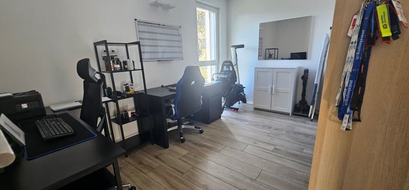 An office setup featuring a black desk with keyboard, computer mouse, and papers. There are two office chairs in front of the desk, and a white wooden cabinet is positioned against the wall. A large window is behind the desk, providing natural light. Abov