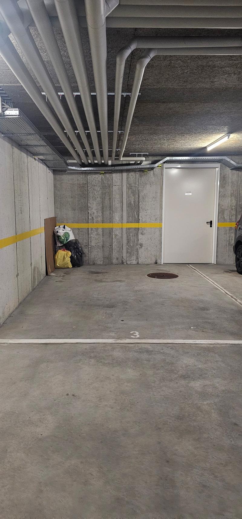 Underground parking lot with concrete walls, yellow and white markings, ceiling with pipes and lights, door on the right side, and some objects like bags near the wall.