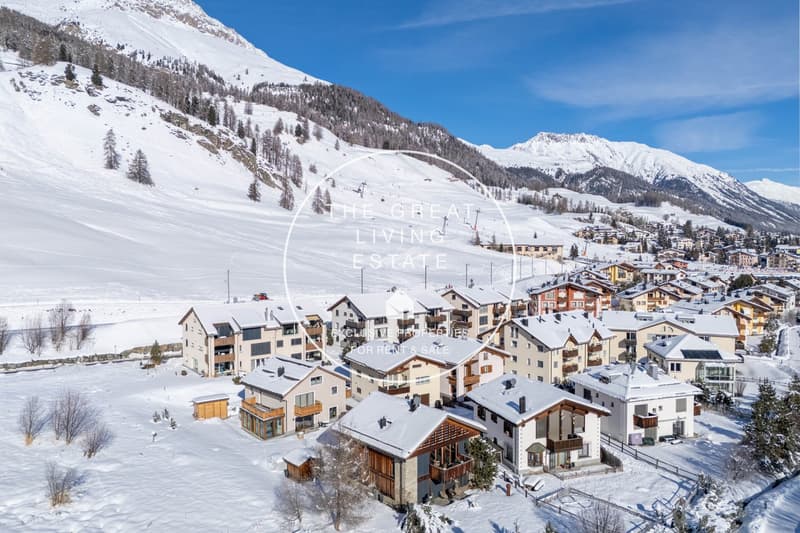 Snowy mountain landscape with a ski resort town nestled in the valley. The town features multi-story apartment buildings and chalets with balconies and parking spaces. The surrounding mountains are covered in snow, creating a picturesque winter scene.