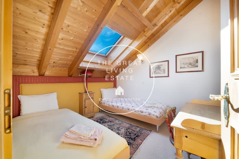 Cozy attic bedroom with wooden beamed ceiling, large skylight, floral patterned rug, wooden furniture, and framed artwork on the walls.