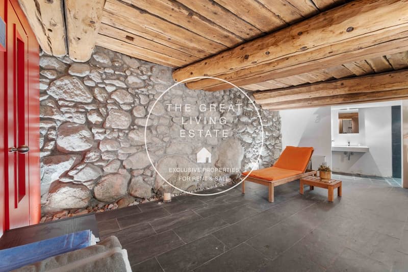 Stone wall, wooden beams, dark flooring, orange lounge chair, small table, candle, and red door. Opposite there's a sink, and further in there's a toilet.