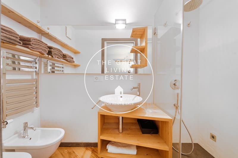 wooden floor, white wall, white sink, glass door, silver faucets, mirror, shelves with towels, small shelf below the sink