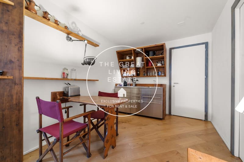 A wooden dining table and two director chairs, a mounted shelf with kitchen utensils, a white door, a kitchen with wooden shelves and a silver fridge, wooden walls.