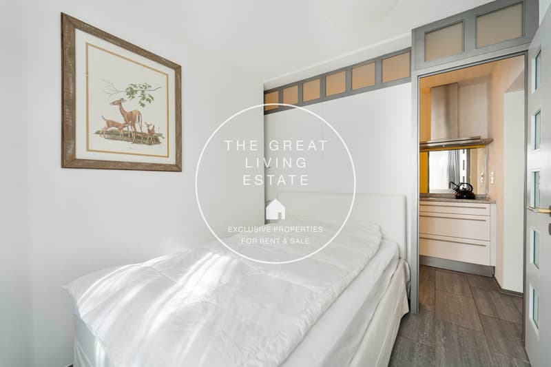 small bedroom, white walls, wooden floor, bed, artwork on wall, open door to kitchen, cabinetry, counter, kettle