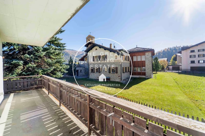 a stone facade house with a tower in front, a wooden balcony fence, large green lawn, trees, mountains in the background