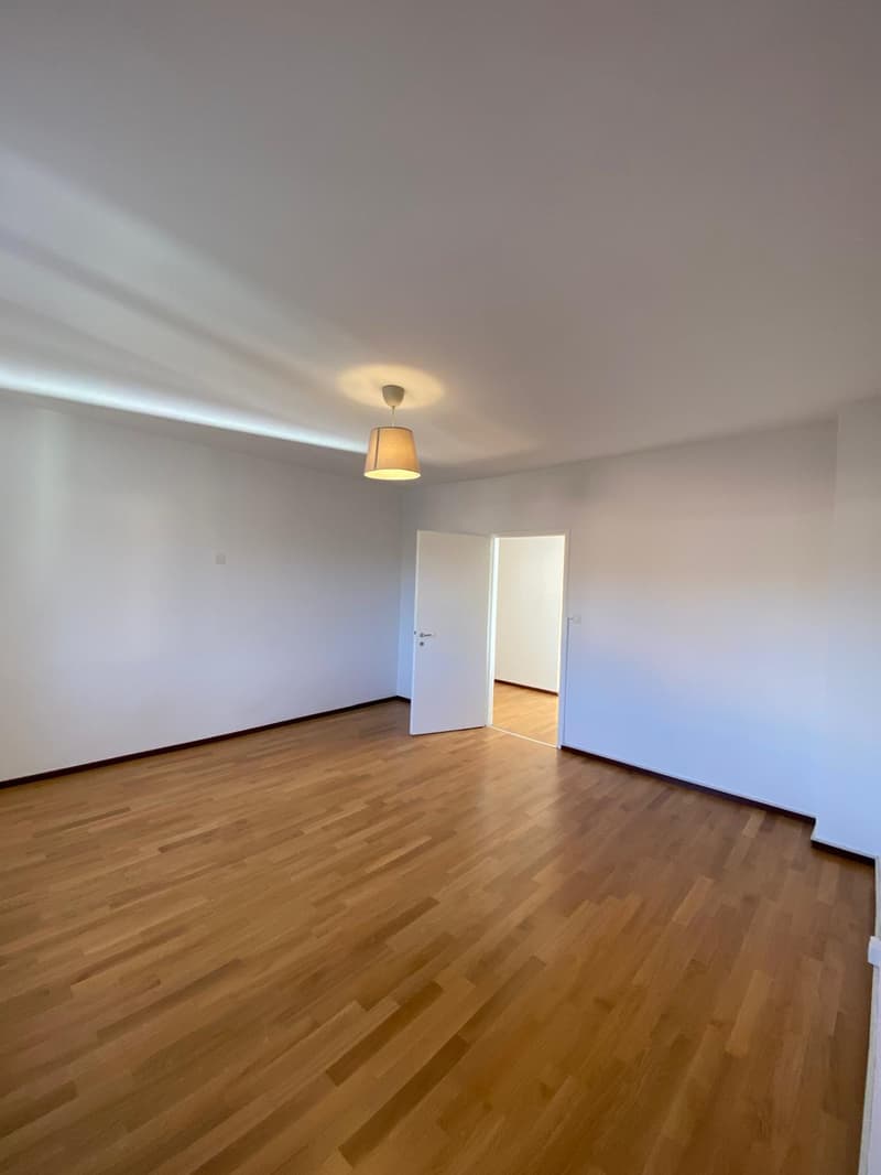 empty room with hardwood floors, white walls, white door, and an open doorway to another room with light on
