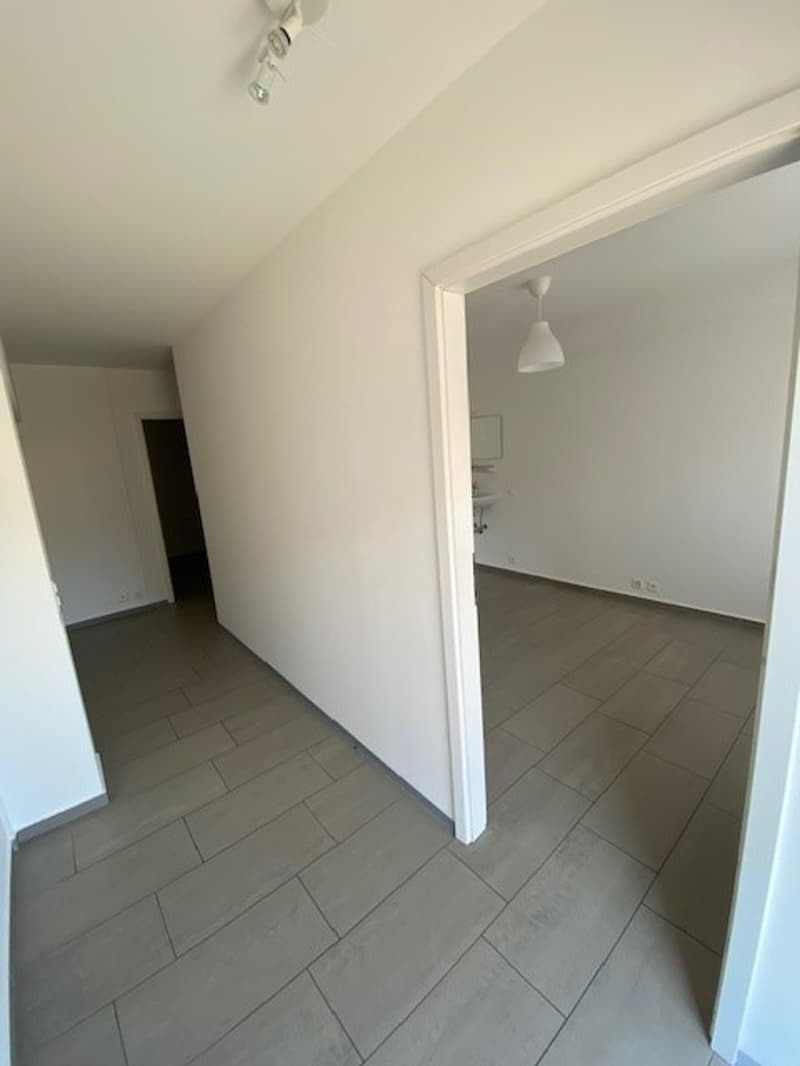 The image shows a hallway with white walls and gray tile flooring. The hallway leads to another room that is visible through the open doorway.