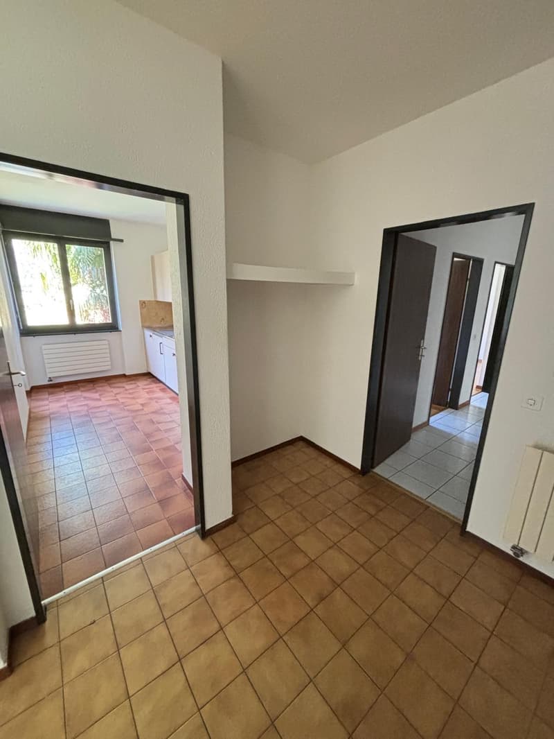 empty room with tiled flooring, white walls, open door, kitchen with window and white cabinets, radiator, hallway