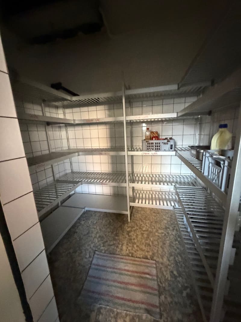 empty storage room with metal shelves and tiled walls, small white bucket on top shelf, small rug on floor