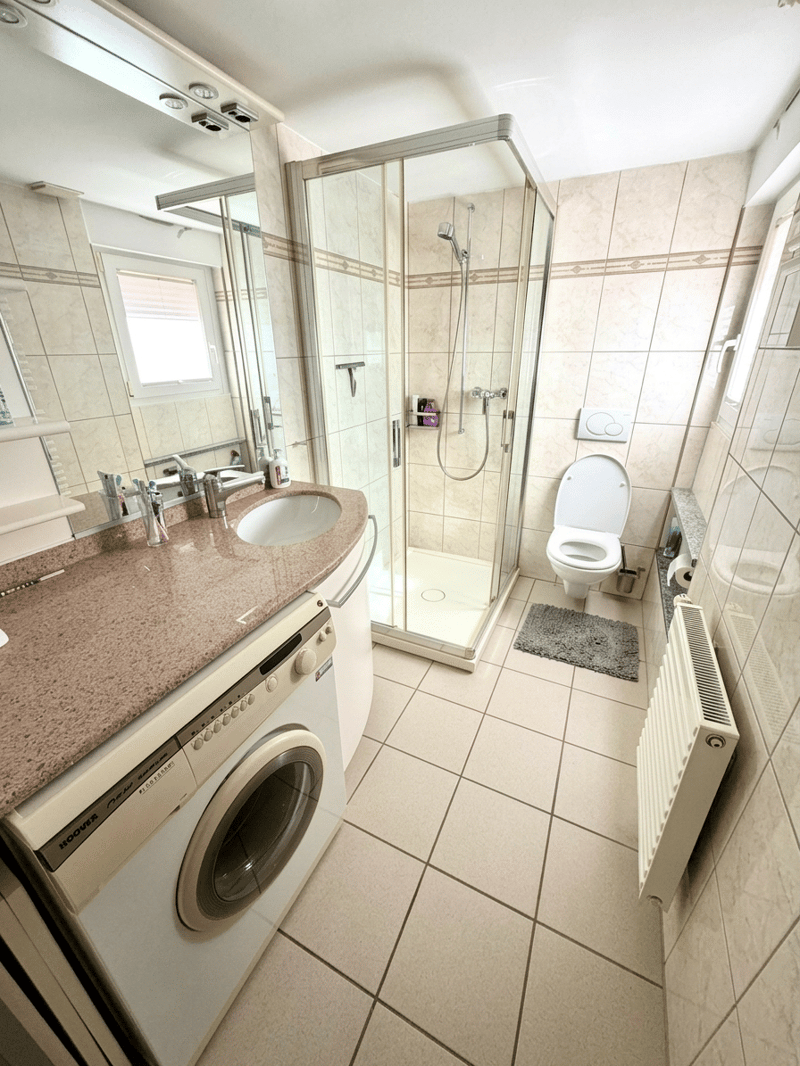 Bathroom with shower, toilet, sink, washing machine, radiator, and tiled floor.