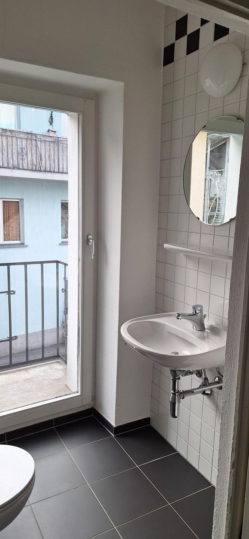 white sink, white toilet, white tiles, black tiled floor, mirror, balcony with railing, open door