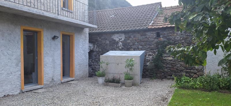 A two story house with a grey exterior, balconies, two entrances, stone walls, gravel courtyard, and plants