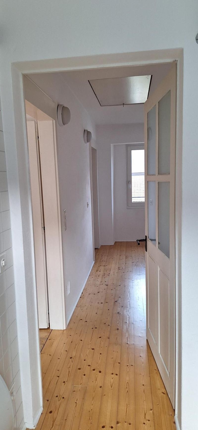 Hallway with hardwood floors, white walls, two wall-mounted lights, open door to another room, closed door, window on right wall