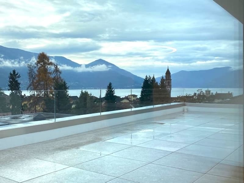 The image shows a balcony or terrace overlooking a scenic landscape. In the distance, there are mountains and a church tower visible. The balcony has a tiled floor and glass railings, providing an unobstructed view of the surrounding area.