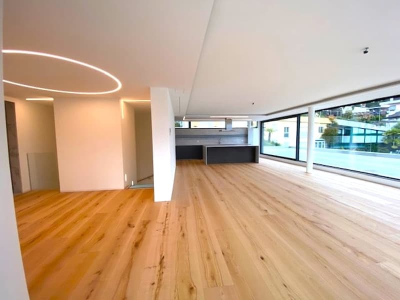 Spacious open-plan living area with hardwood flooring, large windows providing natural light, and a balcony or terrace visible.