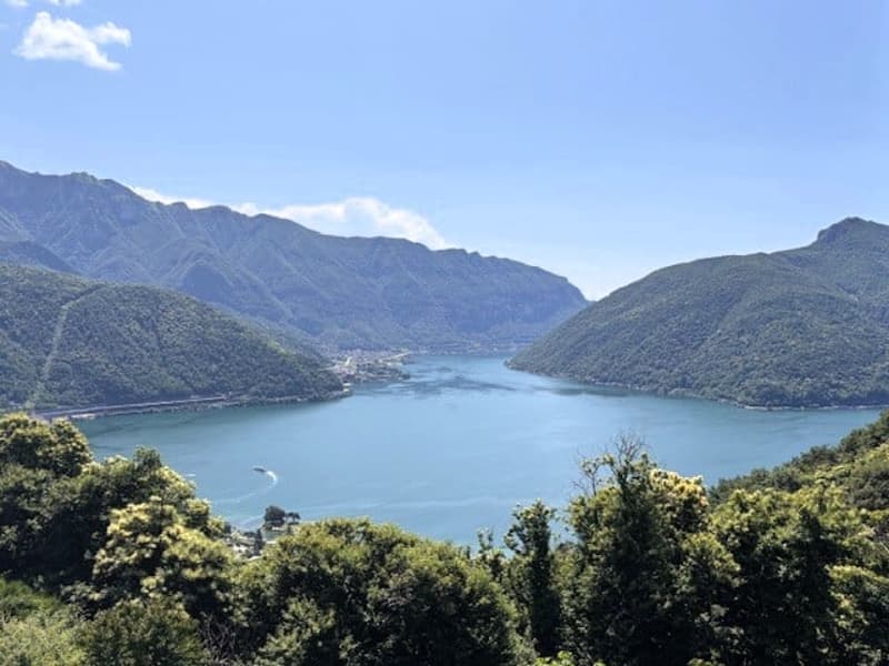The image shows a scenic view of a lake surrounded by mountains and lush greenery. The lake appears to be a deep blue-green color, reflecting the sky and the surrounding landscape. The mountains in the background are covered in dense vegetation, creating