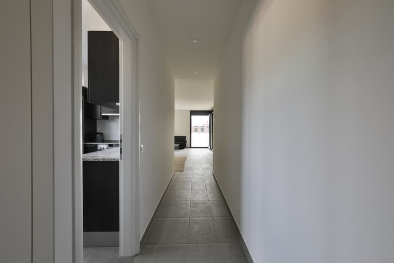 Long, narrow hallway with white walls, gray tile flooring, and a view of a kitchen with black cabinets and a dishwasher visible through an open doorway.
