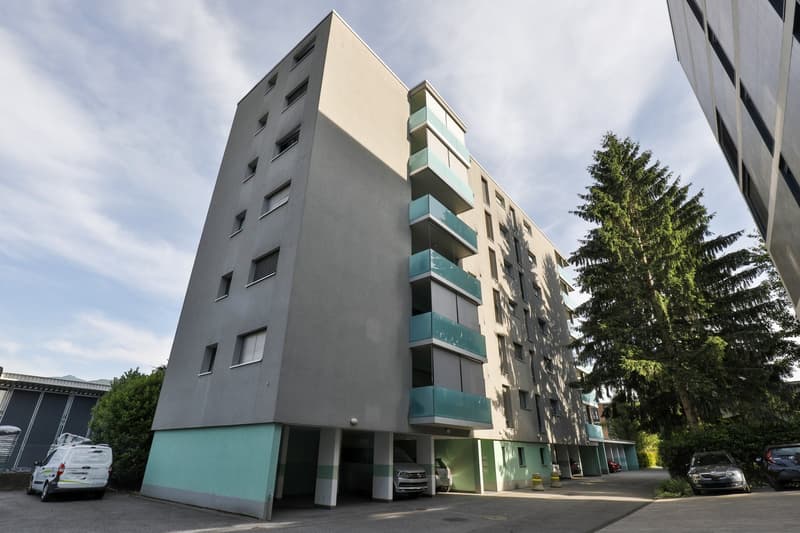 Multi-story apartment building with gray exterior and green accents, balconies, and a parking area in the front
