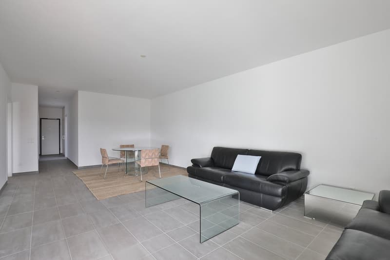 Spacious open-plan living area with grey tile flooring, white walls, black leather sofa, glass coffee table, and a dining table with chairs. The room appears to have a modern and minimalist design.