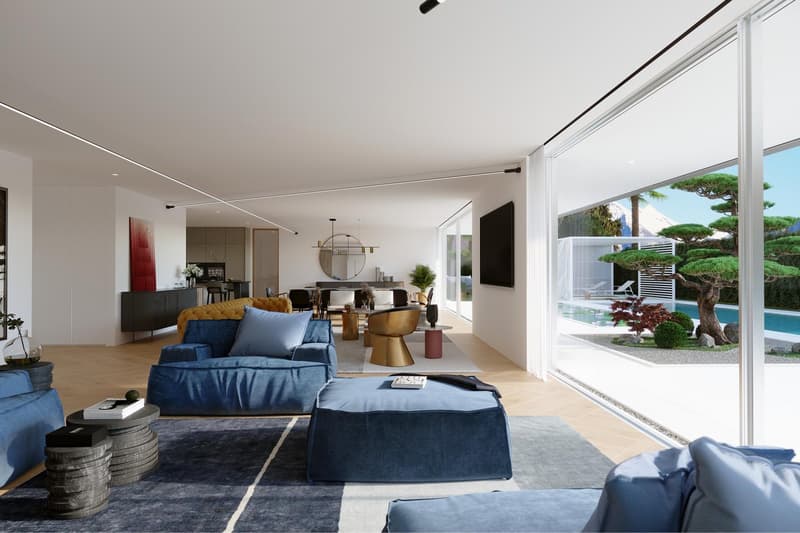 Spacious open-concept living room with large windows overlooking an outdoor garden and pool area. The room features a blue sectional sofa, a round gold coffee table, and a black entertainment unit. The decor has a modern and minimalist style with a mix of
