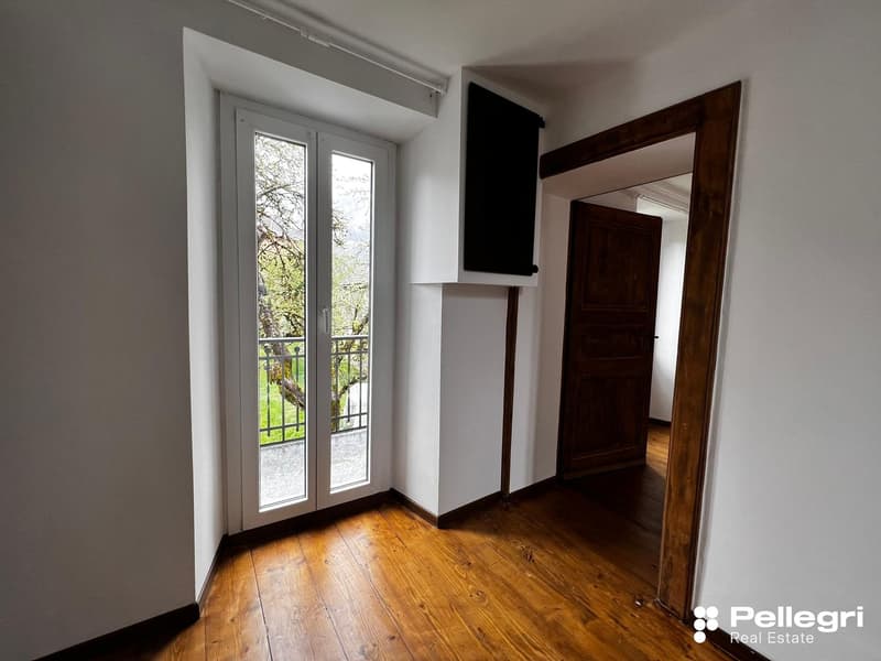 This image shows a room with a large window overlooking a balcony or terrace. The room has white walls and hardwood floors. There is a wooden door frame visible in the background.
