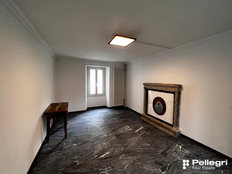 Large room with gray walls, hardwood floor, and a fireplace with a decorative frame. There is a wooden desk and a framed artwork on the wall.
