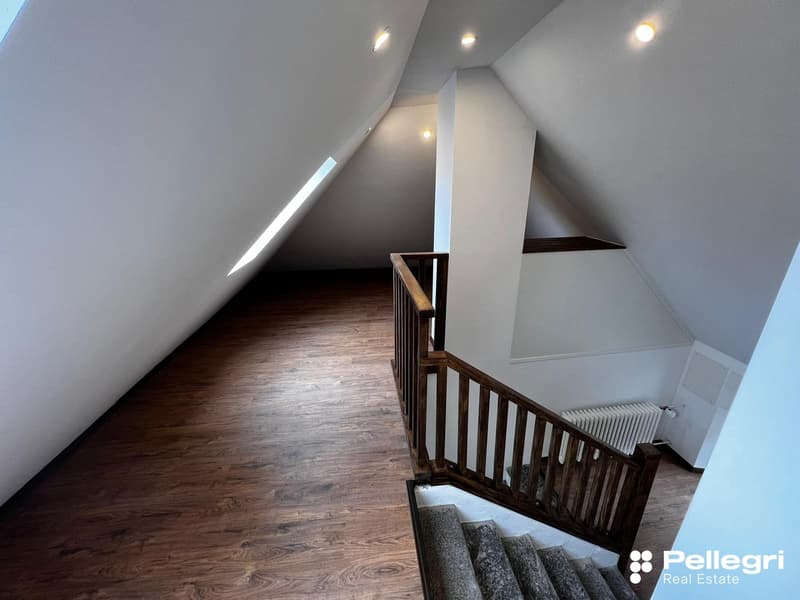 Spacious attic with slanted ceilings, wooden floors, and a wooden railing along the stairs leading up to the space. The room appears to be well-lit with recessed lighting.
