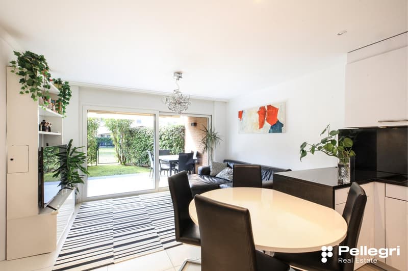 Spacious open-plan living area with large sliding glass doors leading to a balcony or terrace, providing a view of the outdoor greenery. The room features a dining table with chairs, a sofa, and various potted plants. The walls are decorated with artwork,