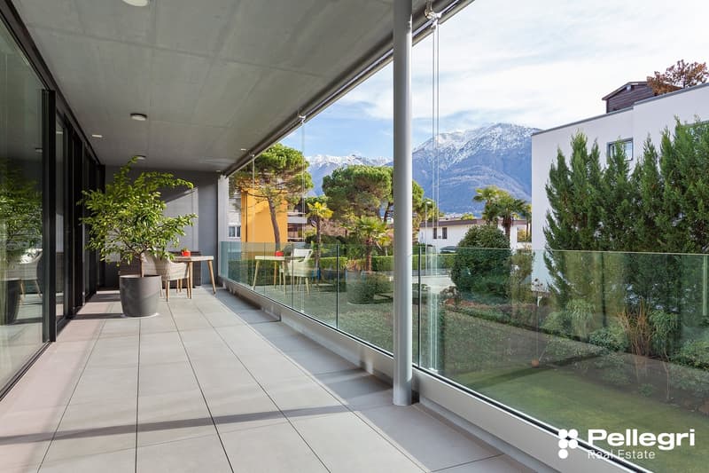 Spacious balcony with glass railing, overlooking a landscaped garden and snow-capped mountains in the background. The apartment features a modern, minimalist design with large windows allowing for abundant natural light and scenic views.