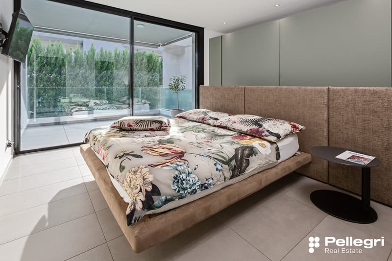 Spacious bedroom with large sliding glass doors leading to a balcony, providing a view of the surrounding greenery. The room features a plush, floral-patterned bedding set, a beige upholstered headboard, and a side table with a lamp.