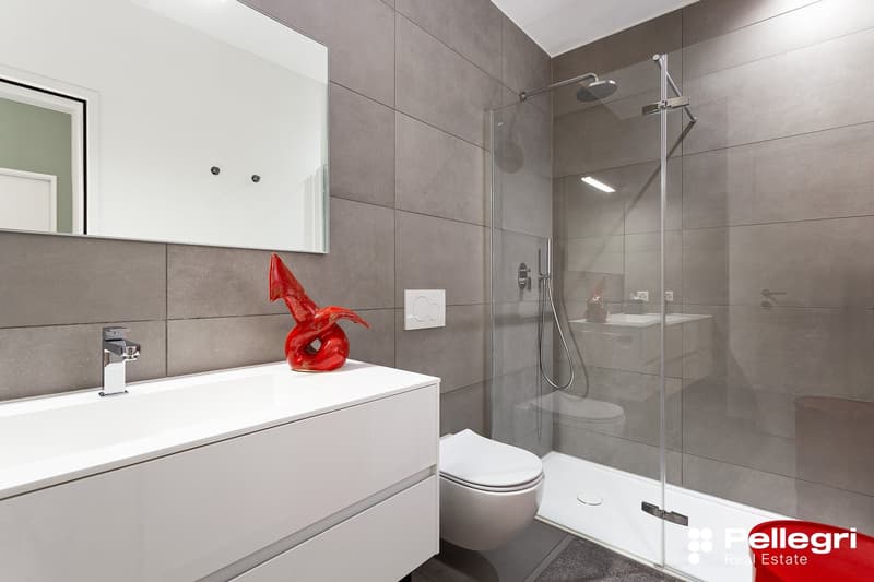 Modern bathroom with gray tile walls, white vanity with sink, red decorative sculpture, glass shower enclosure, and toilet