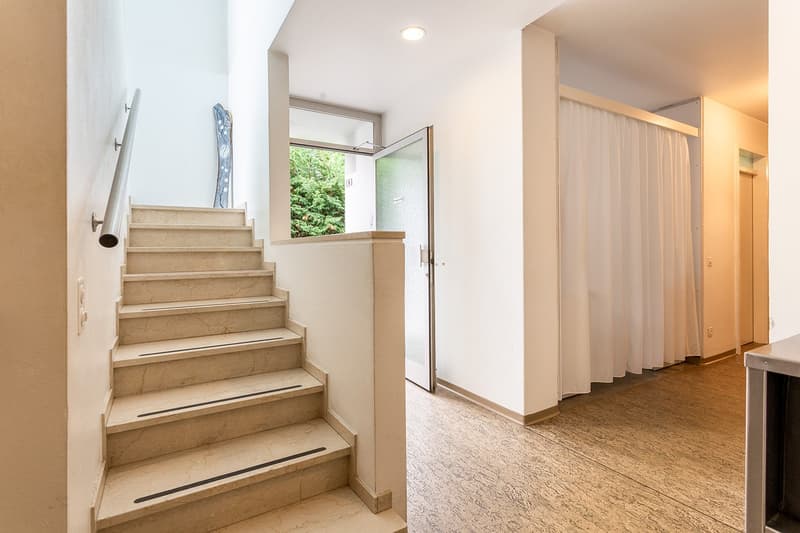 The image shows the interior of a residential property. It features a staircase with beige marble steps leading up to the second floor. The hallway has white walls and a curtain-covered doorway leading to another room. The floor is covered with a light-co